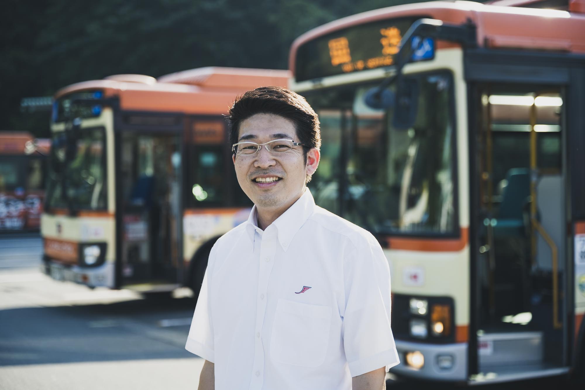 早朝の営業所で、運行表を確認しながら穏やかな表情で今日の業務を把握するバス運転士。制服姿で、背景には朝日に照らされたバスの車体が並ぶ